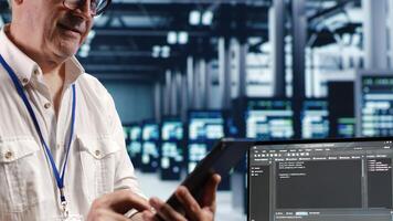 Senior technician in data center, solving tasks on tablet while running scripts on laptop terminal. Licensed serviceman amidst servers, accessing databases and manipulating lines of code photo