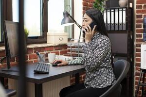 Businesswoman sits at her desk, using her mobile device to talk with a client and sharing updates from her computer. Female employee looking at monitor and having phone conversation with manager. photo