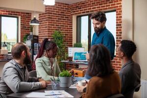 Caucasian manager holding tablet and showing financial graphs to his team seated at table in brick wall office. Bearded businessman using digital device, explaining company growth to his colleagues. photo