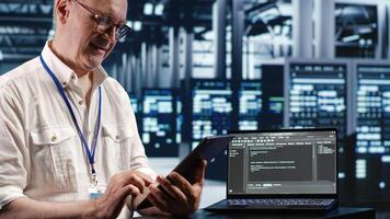 Elderly worker in data center, solving tasks on tablet while running scripts on laptop terminal. Certified engineer amidst servers, accessing databases and manipulating lines of code photo
