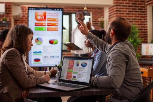 White man raising his hand during meeting with laptop displaying business analytics nearby on table. Selective focus on device screen showing company database research as diverse team have discussion. photo