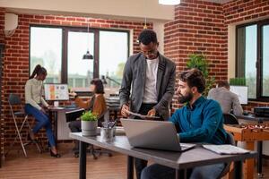 Male professional holds documents and shares ideas with his manager, brainstorming strategies in brick wall workspace. African american analyst with papers in hand, gives feedback to bearded coworker. photo