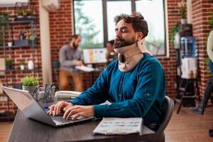 Professional male analyst evaluating research data on a digital device and handling work responsibilities during recovery. Bearded man with a neck brace uses a laptop in a brick-wall office. photo