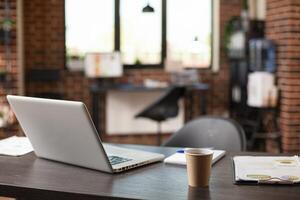 Modern workspace with desk holding digital device, paperwork, and coffee cup, presents professional and organized setup. Vacant table well arranged with office supplies and laptop for employee to use. photo