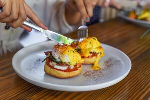 Hands with knife and fork cutting english muffins with poached eggs, bacon and Hollandaise sauce with vegetables photo