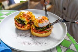 Hands with knife and fork cutting english muffin with poached eggs, bacon and Hollandaise sauce with vegetables photo