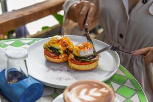 Hands with the knife and fork cutting english muffin with poached eggs, bacon and Hollandaise sauce with vegetables photo