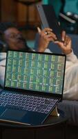 Vertical African american person using his tablet laying on the couch while he pays attention to laptop with stock market data trends. Focused man comparing trading graphs numbers before investing. Camera A. video