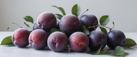 A cluster of fresh plums accompanied by lush green leaves sit on a clean white surface photo