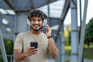 Portrait of a young man on a walk, looking at the camera and smiling, using headphones and an app on his phone to listen to online music, audio books and podcasts. photo