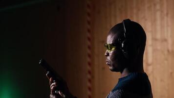 Man in shooting range trying out new gun model before committing to purchase. African american person in indoor firing range learning how to operate new weapon safely, camera B close up video