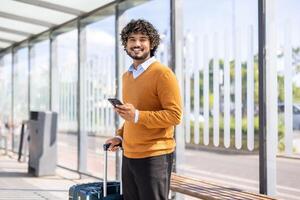 Portrait of a male passenger at a public transport stop, looking at the camera and smiling with a phone in his hands and a white travel suitcase. photo
