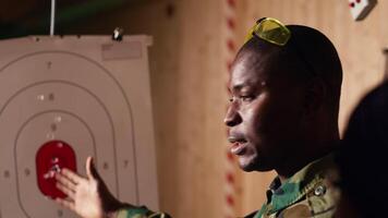 Army leader teaches recruits in shooting gallery how to shoot target center, training accuracy. Military officer instructs soldiers how to hit bullseye, explaining shooting practices, camera B closeup video