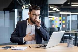 Sick man coughing and having a cold at workplace. Businessman working with laptop at workplace inside office. photo