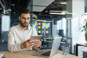 Serious thinking businessman working with tablet computer inside office. Businessman in shirt working at workplace, using laptop, sitting at desk, using financial app. photo