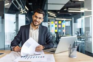 Portrait of successful financier, man signs documents reports contracts. Businessman at workplace inside office doing paper work, filling out tax and statistical forms, looking at camera smiling. photo
