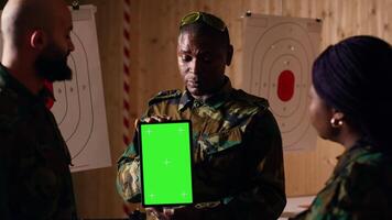 Soldiers at shooting range do training designed to increase safety and survivability in combat, watching instructional videos on green screen chroma key tablet device presented by commander, camera B