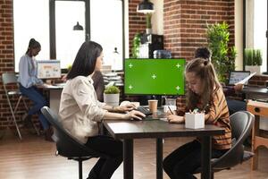 Caucasian mother and daughter sitting at table with computer monitor displaying greenscreen. In startup office, businesswoman using desktop pc with isolated mockup template as little kid sits nearby. photo
