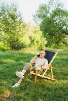 A man is talking on a smartphone while sitting on a bench in a city park. A young guy reads the news, writes a message on his mobile phone. Recreation in nature, remote work on vacation photo