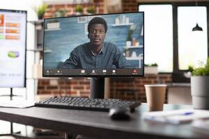 Closeup of computer monitor with virtual meeting in progress on empty desk in startup office. Black man seen on desktop pc in videoconference call as digital screen in background shows analysis data. photo