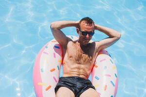 A young man in sunglasses and shorts is relaxing on an inflatable donut in the pool. Summer vacation photo