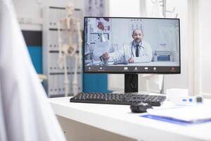 Clinic office desk holds desktop pc with active virtual session showing healthcare specialist. call on computer screen presenting remote medical consultation with caucasian male doctor. photo