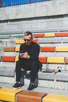 Cheerful hipster guy in trendy outfit smiling while resting at urban setting in city using cellphone device on leisure, happy Middle Eastern male blogger in sunglasses enjoying travel lifestyle photo