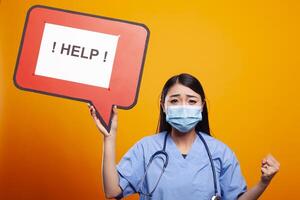 Asian woman doctor with her fist raised, asking for help using a cardboard speech bubble. Female nurse wearing scrubs and face mask, holding a red signboard against an isolated background. photo