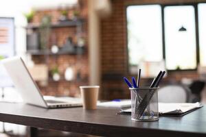 Closeup on container of pens and pencils set on office desk with laptop, cup and documents nearby. Vacant workspace having table neatly arranged with stationery and digital devices, ready for use. photo