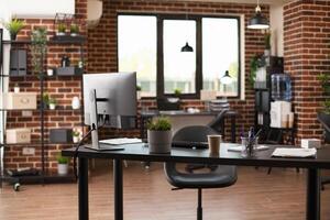 This empty, well arranged workspace has modern desk, desktop monitor, and financial documents. Vacant brick wall room is ready for business projects and marketing strategy development. photo