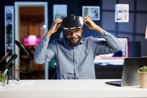 Smiling tech content creator filming technology review of newly released VR glasses, trying them on his head and presenting specifications to audience. Internet star testing virtual reality device photo