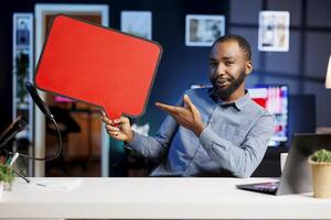 BIPOC man filming sponsored content, holding empty copy space sign in hand, following given script while talking to audience. Online star uses cardboard bubble speech cutout to do promotion to viewers photo