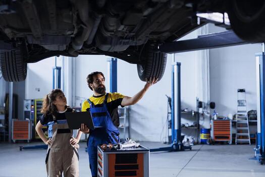 Engineer in car service uses laptop to calculate invoice after fixing broken automobile wheels. Garage worker uses device to inform client of final costs after mending vehicle on overhead lift photo