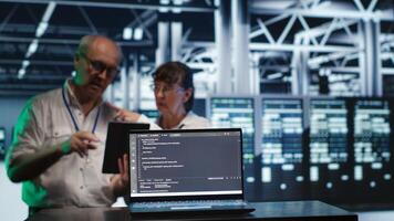 Octogenarian managers in data center, solving tasks on tablet while running scripts on laptop terminal. Adept supervisors amidst servers, accessing databases and manipulating lines of code photo