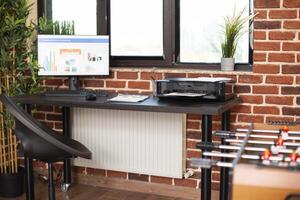 Modern startup office with financial charts on computer screen, printer, and clipboard on table. Bright brick wall workspace with plant decoration and neatly arranged furniture by the window. photo