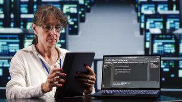 Elderly woman working in IT in high tech workplace completing tasks on tablet while running scripts on laptop terminal. Professional amidst server cabinets manipulates code and improves cybersecurity photo