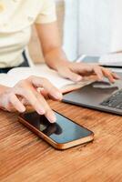 Close-up of hands using a smartphone and laptop on a wooden table, with an open notebook in the background. photo