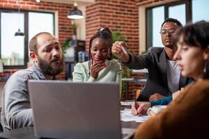 Small group of employees collaborating around a laptop, displaying teamwork in modern office. African american and caucasian colleagues look at device screen, focused on startup project discussions. photo