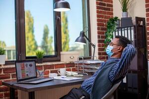Professional analyst with face mask, leaning back in chair and unwinding in brick wall workspace. Businessman looking at laptop screen with hands behind his head, enjoying a break in startup office. photo