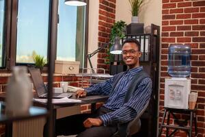 Portrait of black male analyst sitting at neatly arranged desk, looking directly at camera with confidence. Professional man poses with cheerful smile in office, having laptop and documents nearby. photo