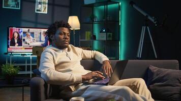 Young adult working remotely from his loft apartment on a laptop. Black freelancer focusing on tasks, blending productivity and relaxation while browsing the web from a cozy sofa. Camera B. video