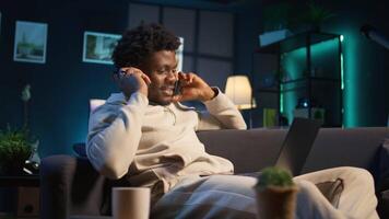 Freelancer listening to a podcast and being focused on online work, putting on headphones while seated on the couch in his living room. Productivity in a casual and flexible home. Camera B. video