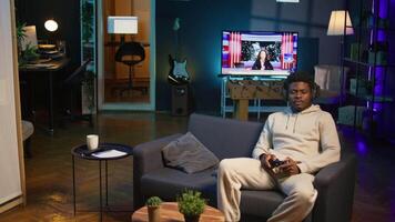 Cheerful man in his apartment playing games on couch, enjoying day off from work. Black gamer battling enemies in online multiplayer shooter on gaming console. Camera B. video