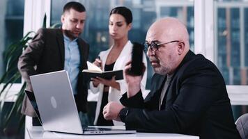 Three diverse business partners having a discussion, working on laptop computer, using mobile phone during meeting at office with large windows showcasing a city skyline. Working together concept video