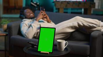 African american young adult laying on the sofa next to mockup copy space, relaxing and browsing web on his smartphone. Happy relaxed person on the couch feels happy with remote job. Camera A. video