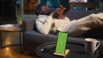 Black male remote worker laying on the couch next to chroma key display, enjoying leisure and free time for relaxation in his modern loft apartment with neon lights and a smart tv. Camera B. video