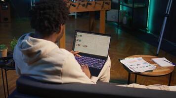 African american person working from home and checking statistical data on laptop screen, analyzing financial graphs. Teleworker in the living room comparing business revenue numbers. Camera B. video