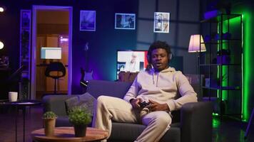 Bored guy plays online game and spamming attack buttons on his controller, modern loft apartment. Gamer in neon lit space participating in internet e sports competition. Camera B. video