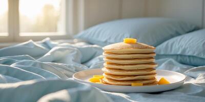 Stack of fluffy pancakes on a bed with light blue bedding and natural sunlight coming through a window with copy space photo