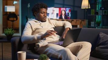 Positive african american man multitasking on a laptop in stylish loft, fully connected to the internet while managing his freelance work and remote responsibilities. Browsing web at home. Camera B. video